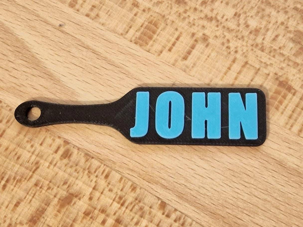 May include: A black paddle-shaped tag with the name "JOHN" in bright blue letters. The tag has a hole at the end of the handle. The tag is laying on a light brown wooden surface. (etsy image id: 7629239329)