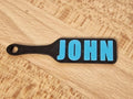 May include: A black paddle-shaped tag with the name "JOHN" in bright blue letters. The tag has a hole at the end of the handle. The tag is laying on a light brown wooden surface. (etsy image id: 7629239329)