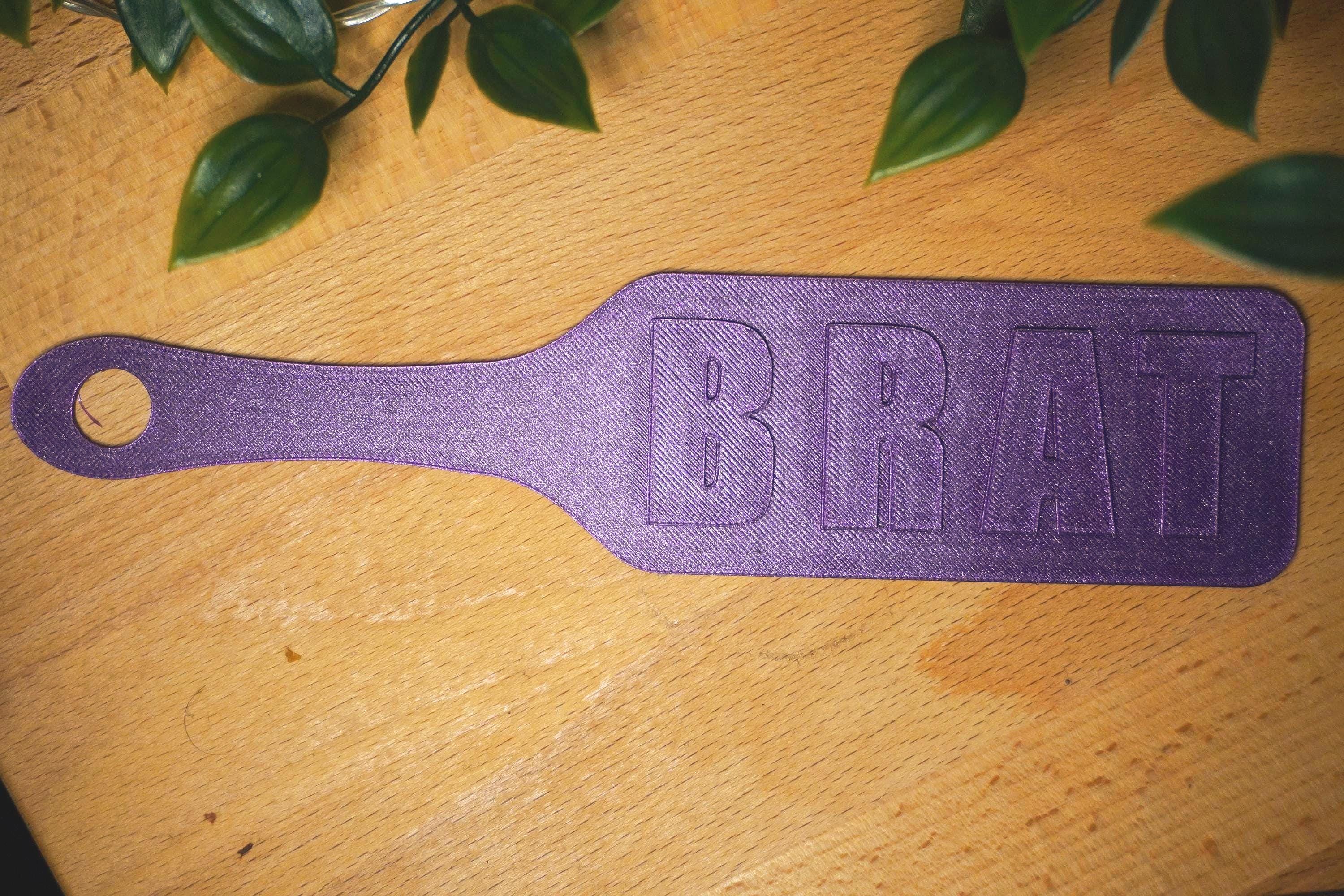 May include: A purple paddle with the word "BRAT" embossed on the surface. The paddle has a handle with a hole at the end. The item is on a wooden surface with green leaves in the background. (etsy image id: 7957328396)