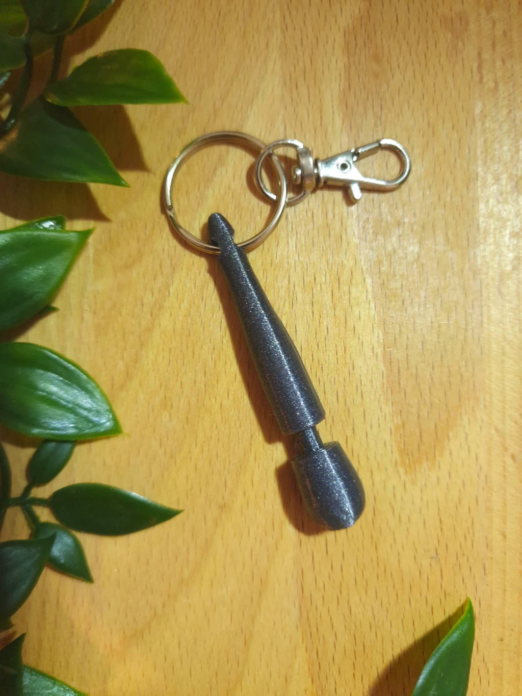 May include: A dark grey, 3D-printed keychain with an elongated, stylized design and a rounded end. It has a silver-coloured split ring and a lobster clasp for attaching to bags or keys. (etsy image id: 7957336362)