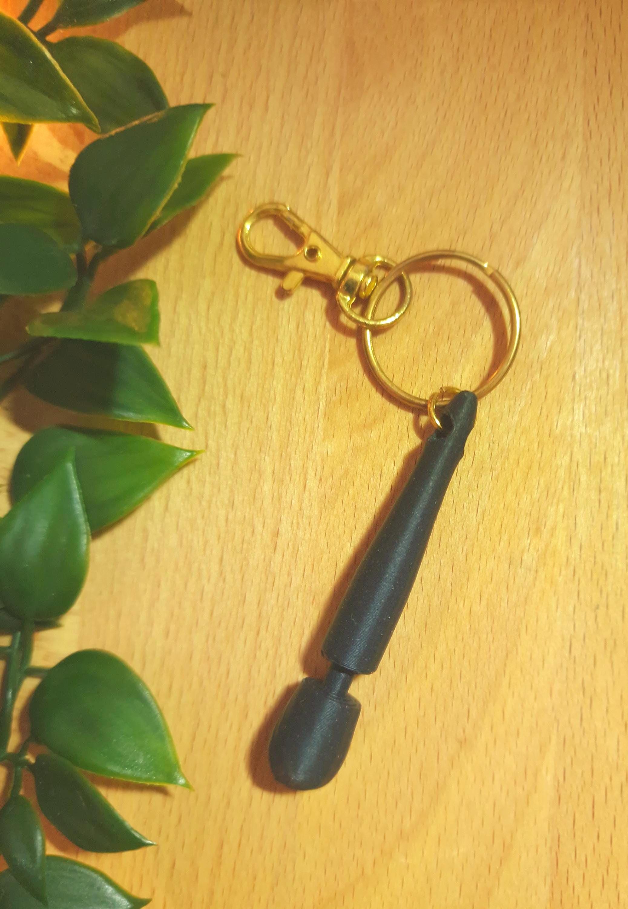 May include: A black keychain with a gold-coloured clasp and ring. The keychain has a black, tapered, cylindrical design. The background is a light brown wooden surface with green leaves. (etsy image id: 8005301381)